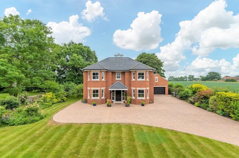 Aerial view of a large red brick house with manicured lawn and driveway, surrounded by green trees and countryside landscape