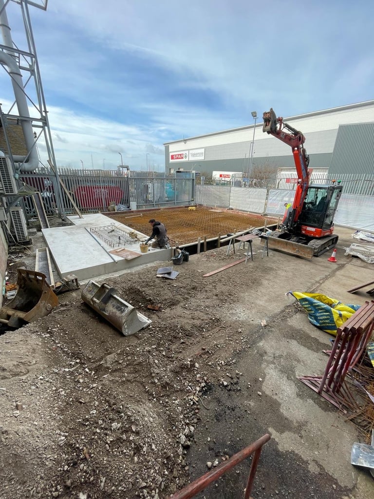 Construction site with worker laying foundation boards on dirt ground, red excavator nearby, industrial building and fencing in background