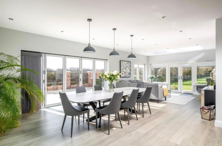 Modern dining room with white table, black chairs, pendant lights, and large windows overlooking a garden