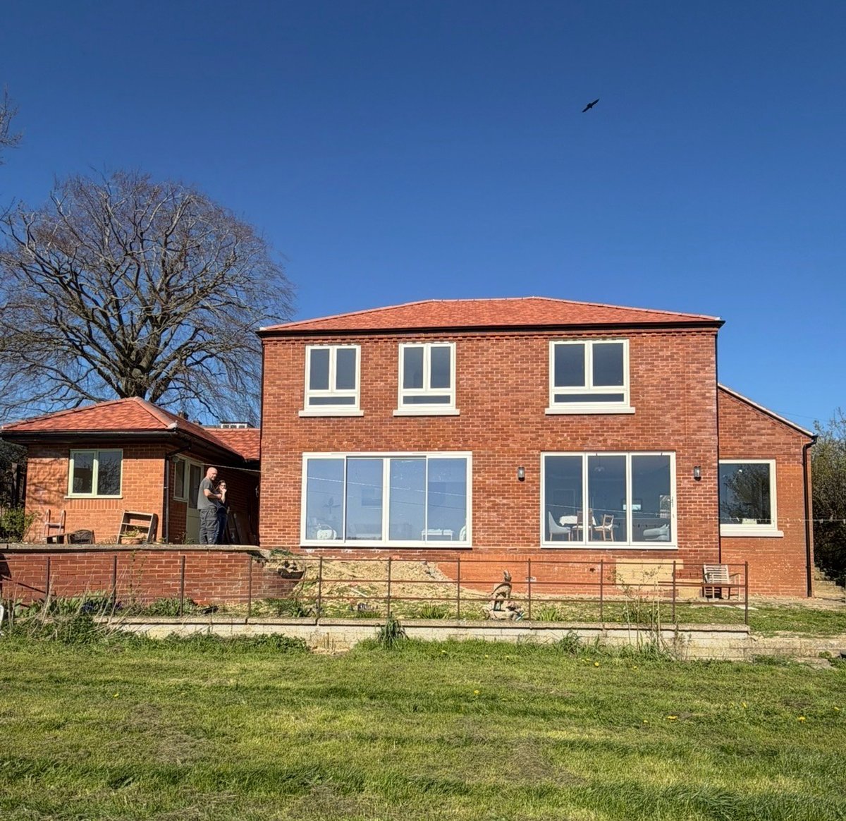 Two-story red brick house with large windows and metal fence, set on a grassy lawn with bare tree and clear blue sky