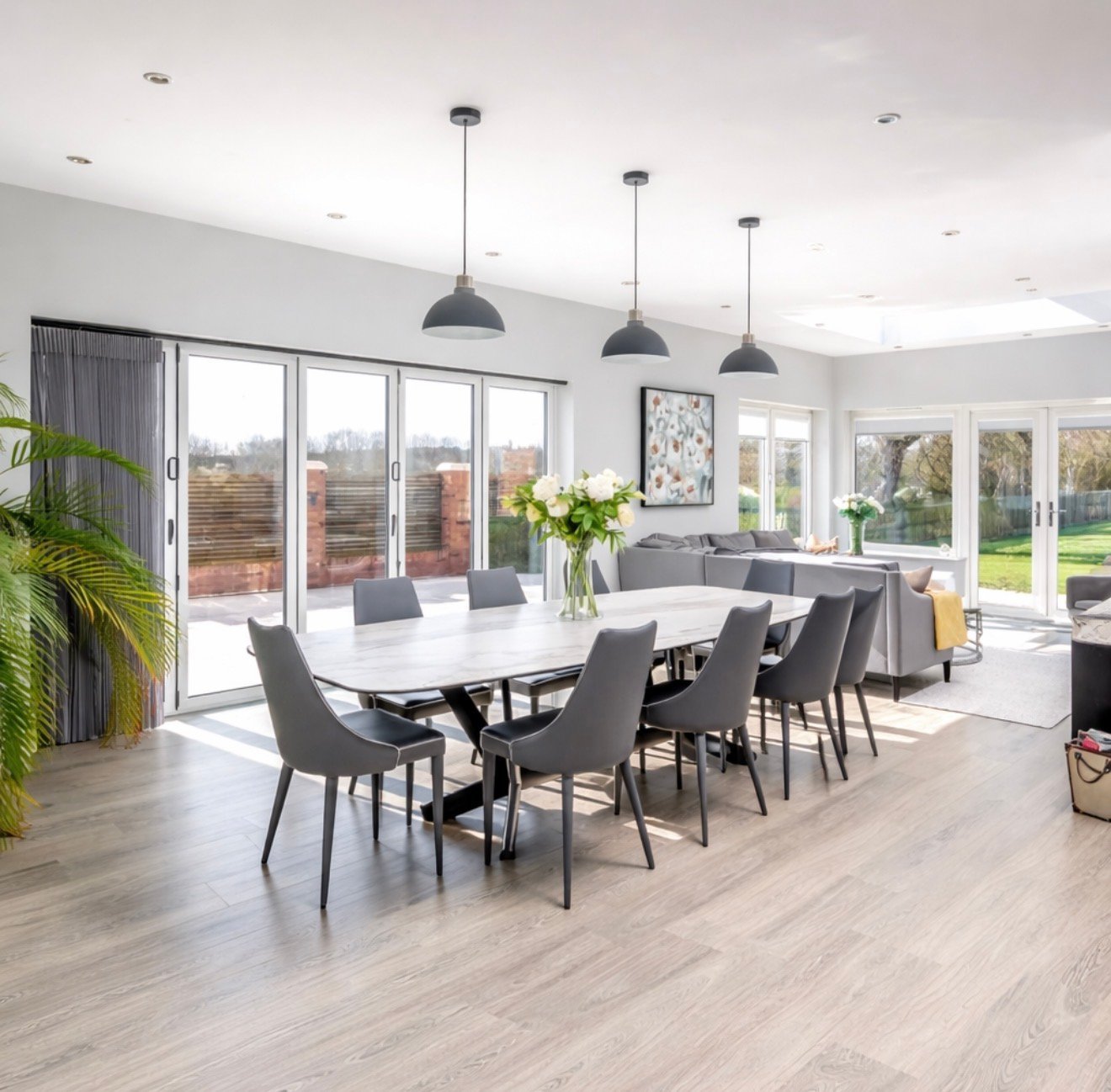 Modern open-plan dining and living room with white marble table, black chairs, pendant lights, and glass doors overlooking garden