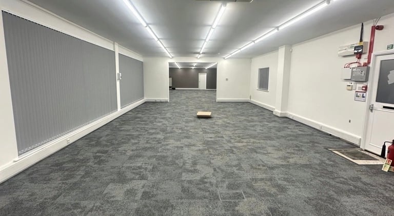 Empty commercial office or retail space with gray carpet flooring, white walls, fluorescent ceiling lights, and utility boxes on the right wall