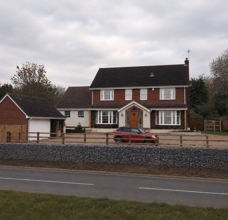Red brick two-story house with white trim and wooden fence, parked red car in driveway, surrounded by trees