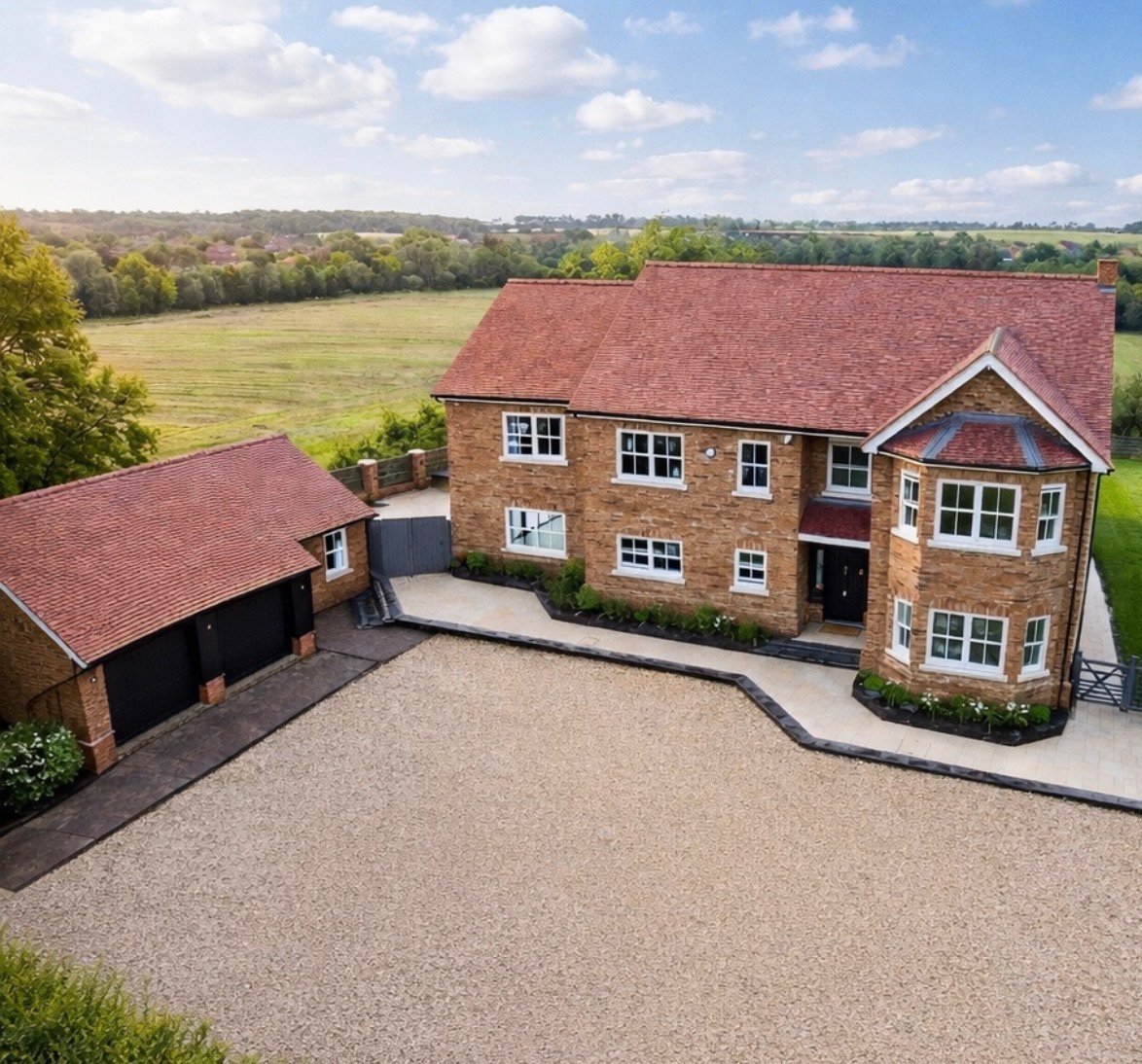 Aerial view of a large brick house with red tiled roof, driveway, and detached garage overlooking green countryside