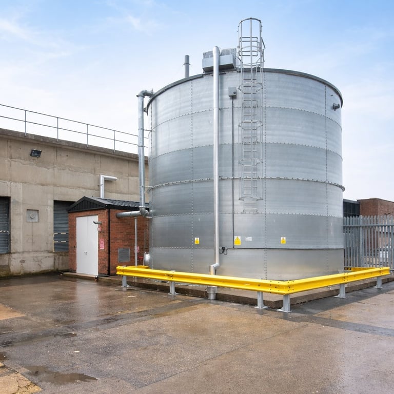 Large cylindrical metal storage tank with yellow safety barriers and climbing ladder at industrial facility